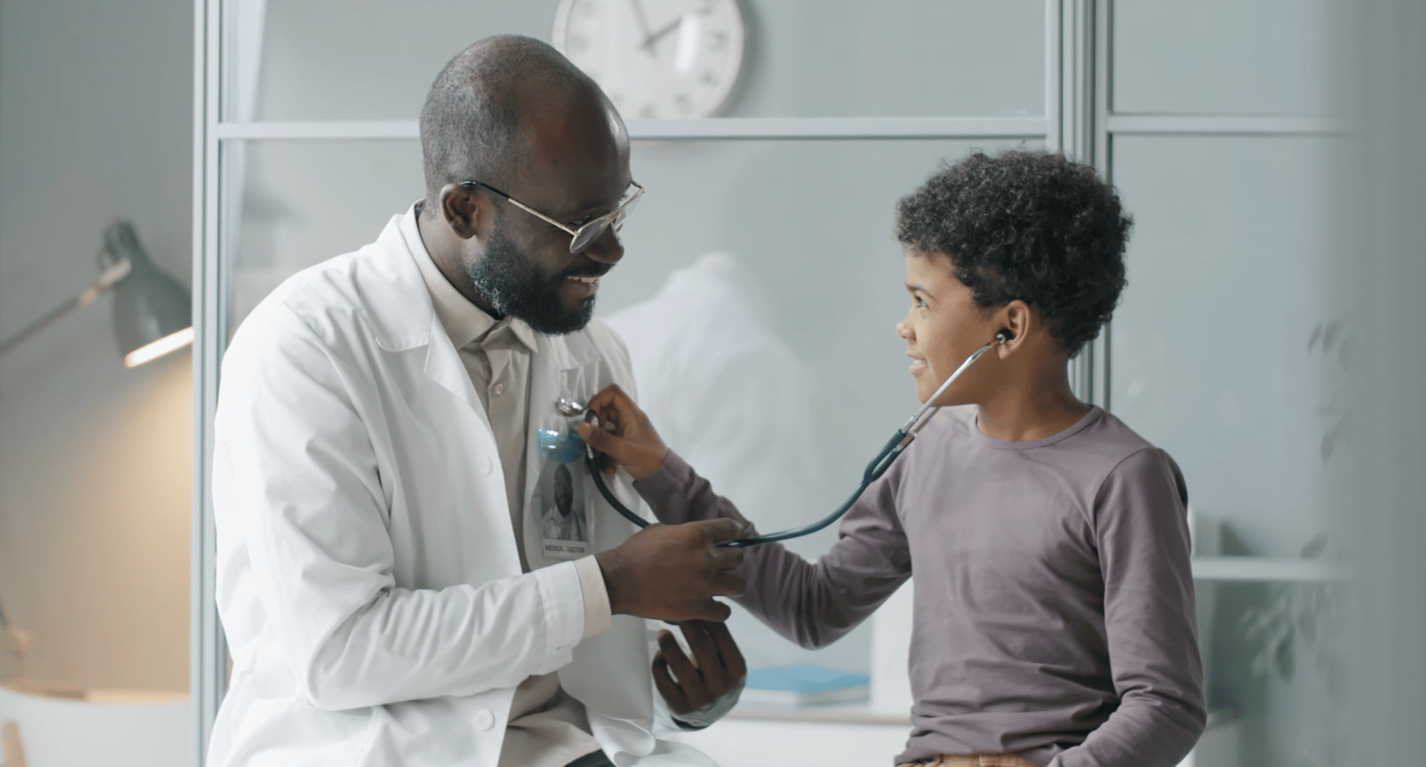 A smiling doctor in a white coat lets a young child playfully listen to his heartbeat with a stethoscope in a bright medical office.