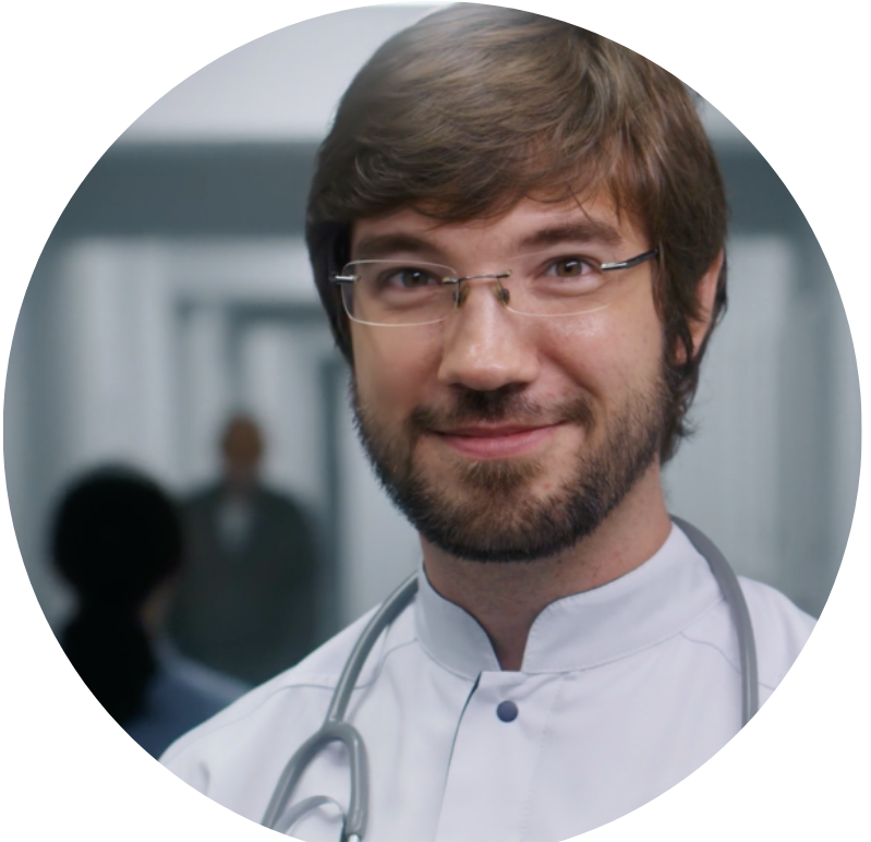 A smiling doctor with glasses and a beard wears a white coat and stethoscope, standing in a brightly lit hospital corridor.