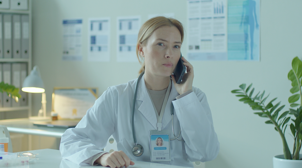 A female doctor in a white coat with a stethoscope around her neck is on the phone, appearing attentive and focused. She sits at a desk in a clinical office setting with medical charts and certificates on the wall in the background.