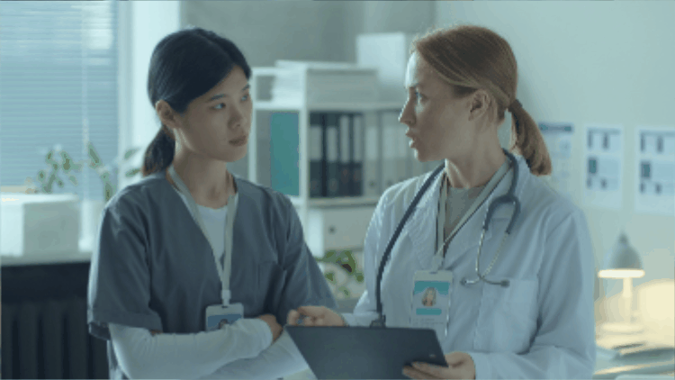 Two healthcare professionals in discussion: a female doctor in a white coat and stethoscope consults with a nurse wearing gray scrubs. They appear focused and engaged in a conversation within a clinical setting.