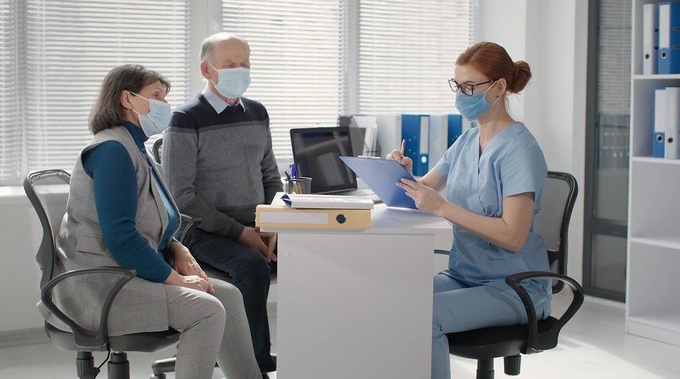 A healthcare provider in blue scrubs and a mask consults with two senior patients, also wearing masks, in a clinical office. The provider reviews information on a clipboard, engaging attentively with the patients seated across from her.